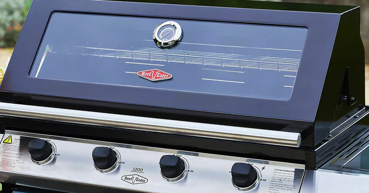 Close-up of a BeefEater 1200 series black and stainless steel gas barbecue with glass lid and four burner knobs.