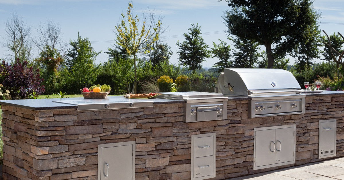 A beautifully designed outdoor kitchen in an autumn garden setting, featuring a built-in BBQ, pizza oven, and stone worktops